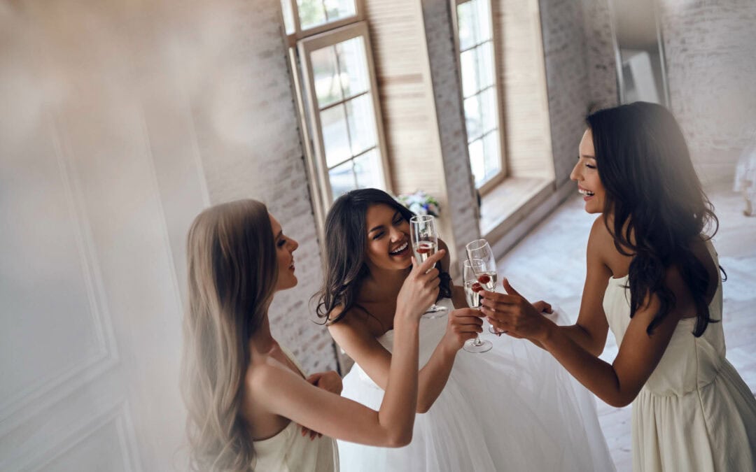 Three women in gowns are smiling and toasting with champagne glasses in a bright, airy room with large windows and brick walls.