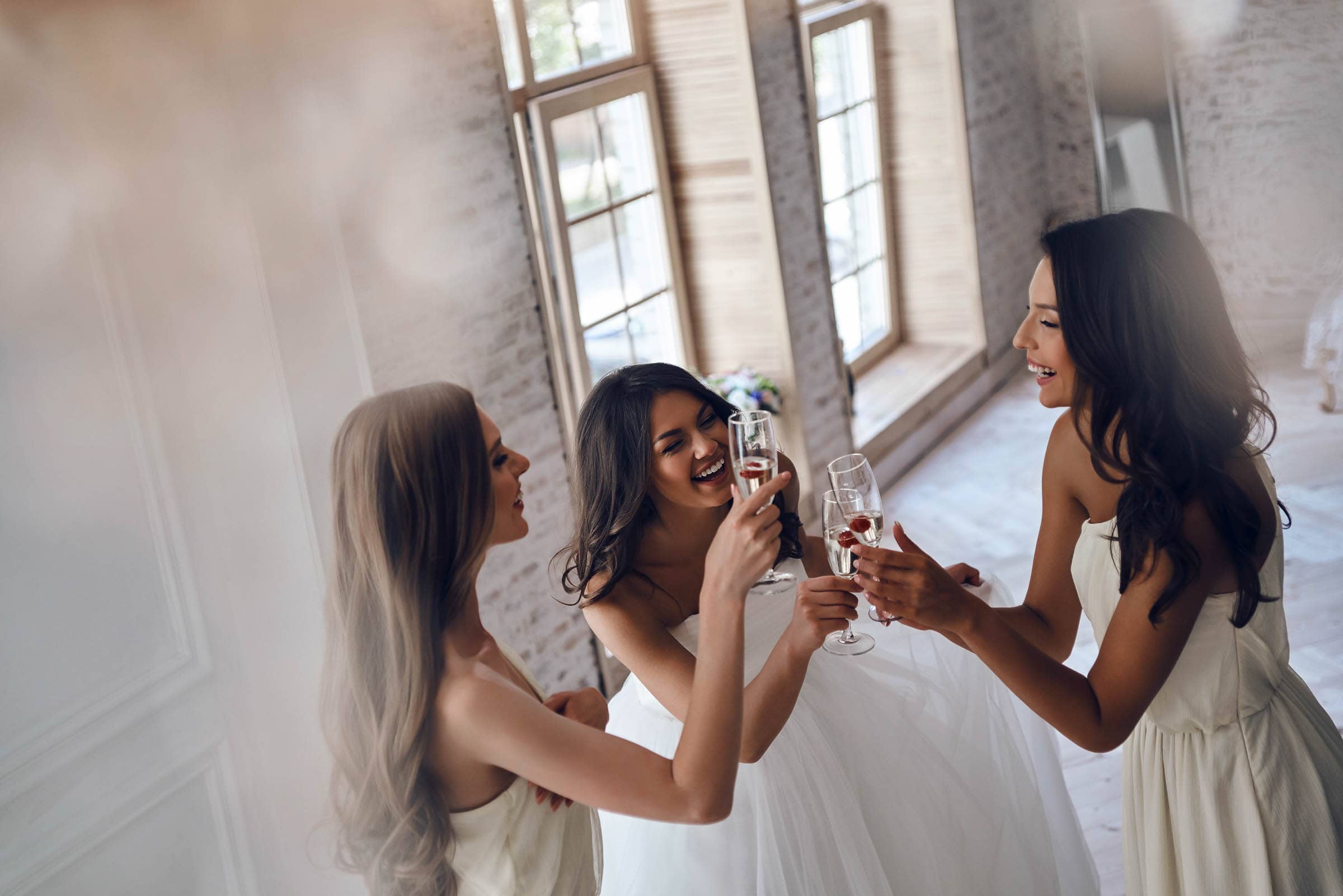 Three women in gowns are smiling and toasting with champagne glasses in a bright, airy room with large windows and brick walls.