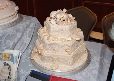 A three-tiered cake with white frosting, intricately decorated with shell-shaped designs. The cake sits on a silver platter on a table.