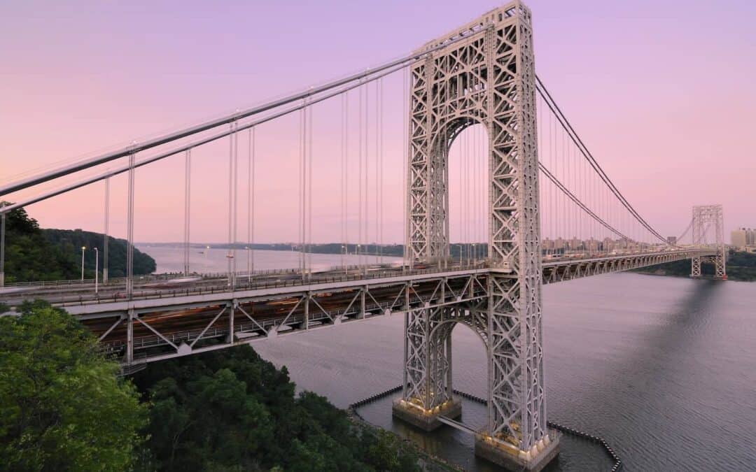 View of the George Washington Bridge spanning the Hudson River with a backdrop of a pink sky at dusk. Trees and water are visible in the foreground and background.