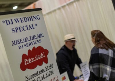 A man in a hat speaks to a woman at a booth advertising a DJ wedding special. The sign lists prices and states Price Slash! Normal rate $1600, Special $1200.