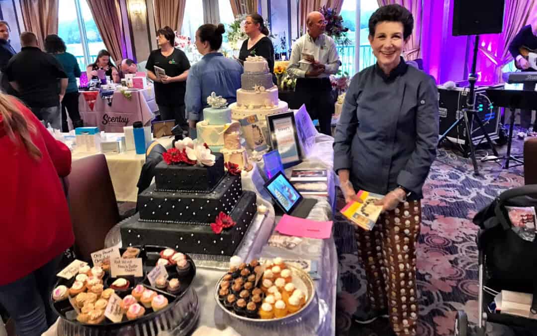 A woman stands by a table with a variety of decorated cakes and cupcakes on display at an expo, with other participants and tables visible.