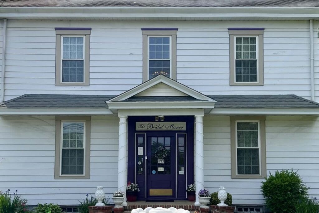 A 2 story white house with a dark blue door under a small porch with 4 windows in front, flower pots, and manicured bushes. The door sign reads The Bridal Manor.