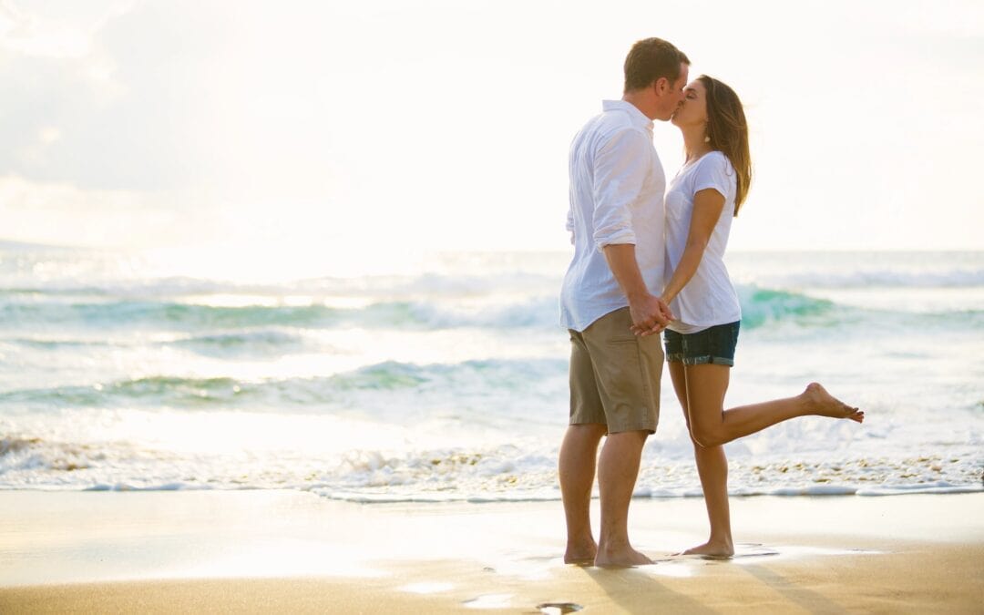 A couple is standing on a beach, kissing, with the woman lifting one leg. They are both wearing casual summer clothes, and the sun is setting in the background.