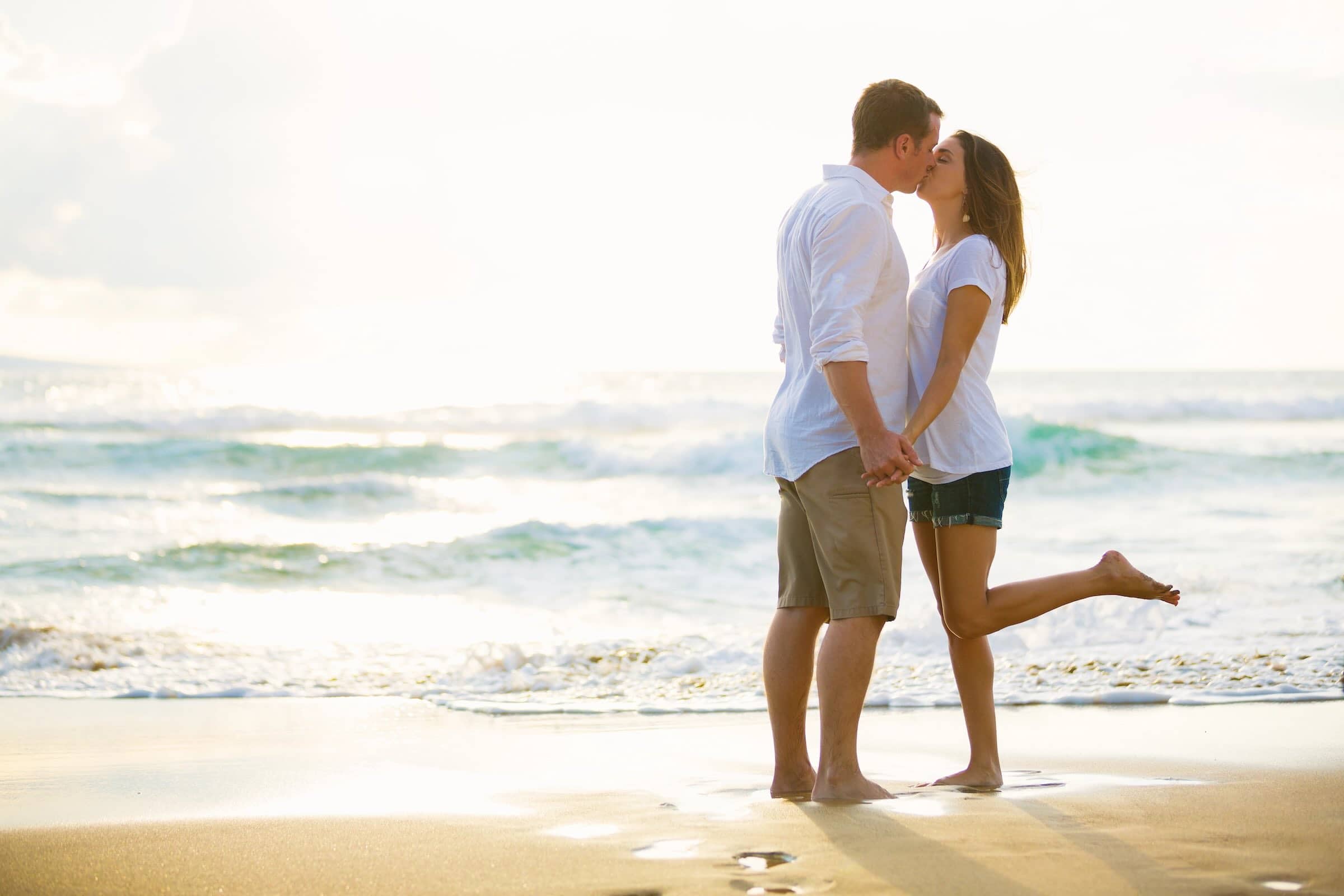 A couple is standing on a beach, kissing, with the woman lifting one leg. They are both wearing casual summer clothes, and the sun is setting in the background.