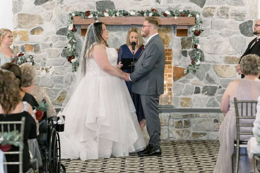 A couple stands at the altar during their wedding ceremony, holding hands and facing each other. The officiant is reading, and guests are seated on both sides.