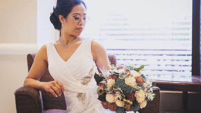 A bride in a white dress and glasses sits on a chair while holding a bouquet, looking to the side.