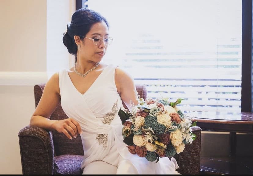 A bride in a white dress and glasses sits on a chair while holding a bouquet, looking to the side.