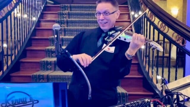 A man in formal attire plays an electric violin on a stage, with audio equipment and a microphone in front of him. A staircase with ornate railings is behind him.