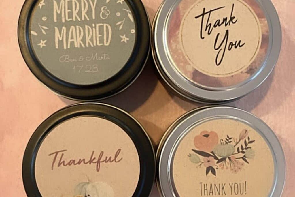 4 round tin containers with decorative labels on a pink surface. The labels read Merry and Married, Thank You, Thankful, and Thank You! with various designs and fonts.