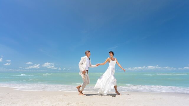 Bride and groom holding hands and walking barefoot on a sandy beach, with the ocean and clear blue sky in the background.