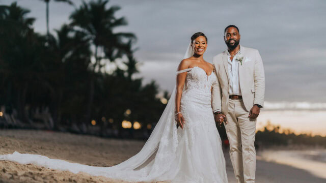 A couple in wedding attire stands on a beach at sunset, with palm trees and a cloudy sky in the background.