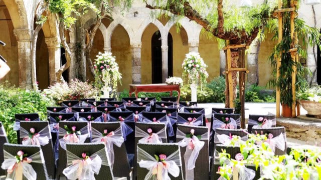 Outdoor wedding ceremony setup with black chairs adorned with white ribbons, in rows. A wood table and flowers are at the front, set against an arched stone background.