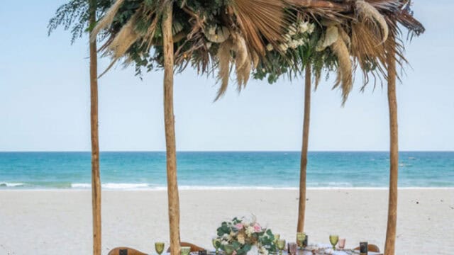 A beach setup featuring a wooden canopy adorned with dried palm leaves and greenery, shading a dining table with chairs on the sand.