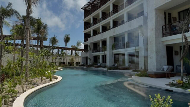 A multi-story resort building with balconies overlooks a large, curved outdoor swimming pool surrounded by lush greenery under a partly cloudy sky.