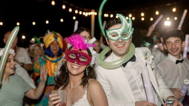 A bride and groom wearing festive masks and surrounded by party guests celebrate under string lights at night.