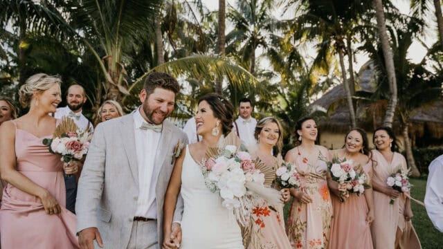 A wedding party, including the newlyweds and bridesmaids in light pink dresses holding bouquets, walk together outdoors with palm trees in the background.