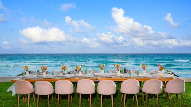 A long table set for a meal with pink chairs, floral centerpieces, and tableware arranged outside on grass, facing the ocean under a blue sky with clouds.