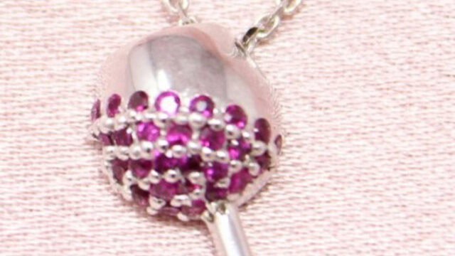 A silver necklace pendant shaped like a wine glass, decorated with small pink gemstones, rests on a light pink fabric.