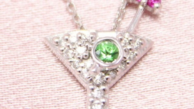 A silver necklace with a pendant shaped like a martini glass encrusted with small diamonds, a green gem in the center, and a pink jeweled umbrella on the side.