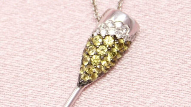 A necklace pendant shaped like a champagne flute, adorned with yellow and clear gemstones, on a light pink background.