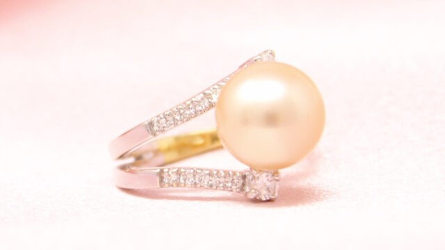 A gold ring with a large pearl centerpiece and small diamonds on the band is photographed against a soft pink background.