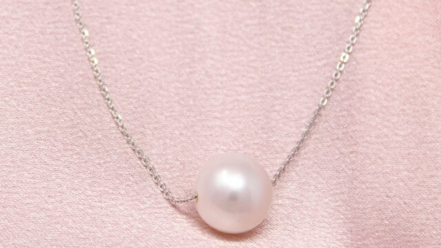 A single pearl necklace on a delicate chain is placed on pink satin fabric.