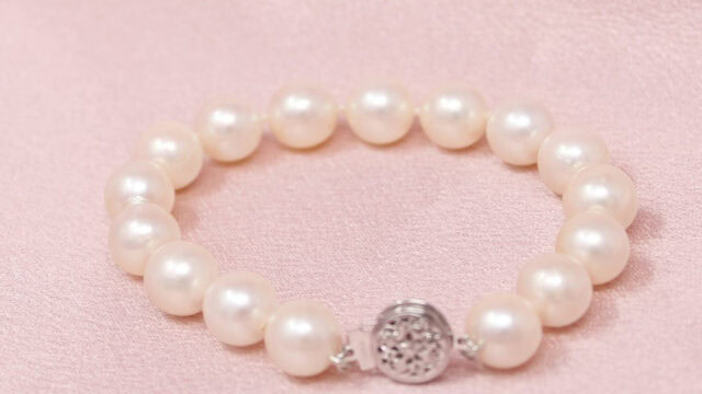 A pearl bracelet with a silver clasp featuring an intricate design, resting on a light pink fabric background.