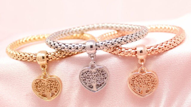 Three metallic bracelets with intricate tree-heart charms, displayed on a soft pink surface. The bracelets are in gold, silver, and rose gold tones.