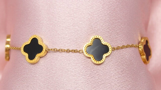 Close-up photo of a gold bracelet featuring black enamel clover-shaped charms, displayed on a pink fabric background.