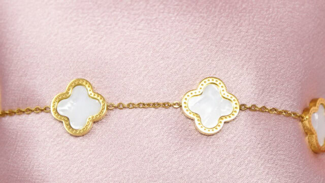 A gold chain bracelet featuring white clover-shaped charms, displayed against a light pink fabric background.