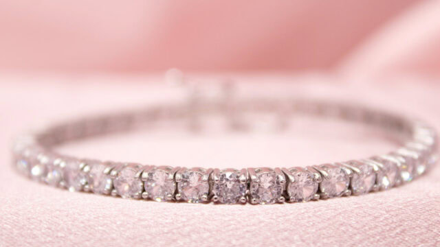 A close-up of a silver tennis bracelet with round, clear gemstones set in prong settings, placed on a soft pink fabric background.