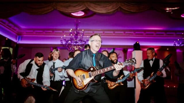 A man in formal attire enthusiastically plays an electric guitar on stage at a wedding reception, surrounded by men in suits holding inflatable guitars.