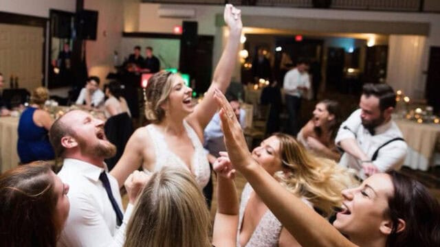 A group of people in formal attire dance and celebrate enthusiastically at an indoor event, likely a wedding reception, under dim lighting.