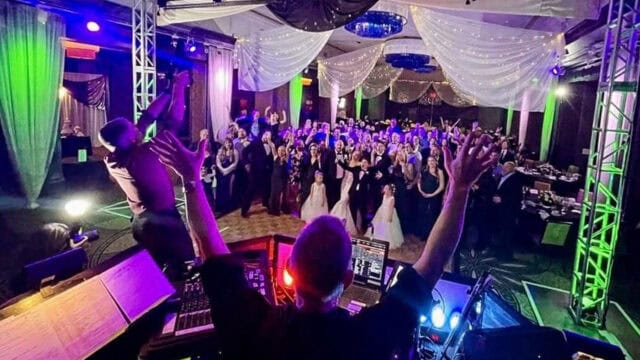 A DJ stands at his equipment with arms raised, facing a crowd of people dancing and celebrating under draped fabric and colorful lights at a wedding reception.