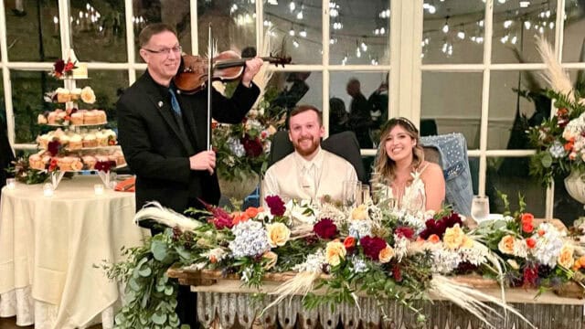 A bride and groom sit at a table with flowers, smiling at the camera. A violinist stands next to them playing the violin. A cake display is visible in the background.