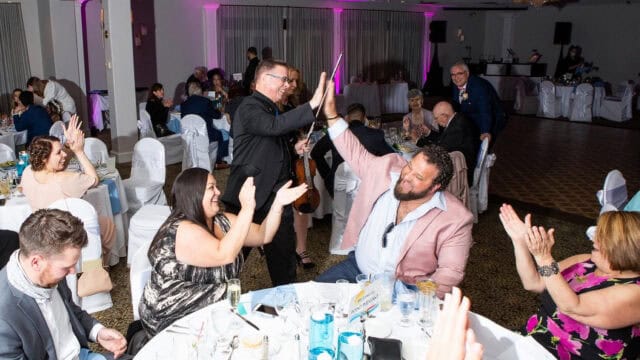 People at a banquet hall cheering and high-fiving around dinner tables, some clapping and smiling, with the room lit by ceiling lights and pink uplighting.