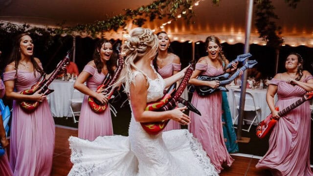 A bride in a white wedding dress dances with bridesmaids in pink dresses holding inflatable guitars under string lights at a reception.