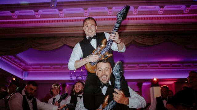 2 men in formal attire are joyfully playing inflatable guitars, with one sitting on the shoulders of the other in a lively, purple-lit room.