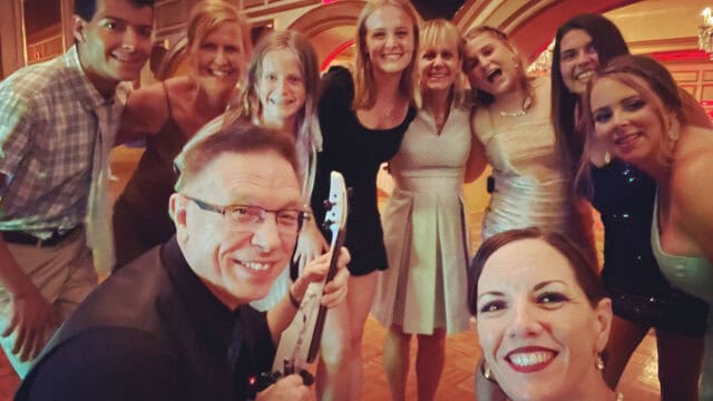 A group of people posing for a selfie at an indoor event. Some are smiling and others are making playful expressions. The setting appears to be a festive gathering.