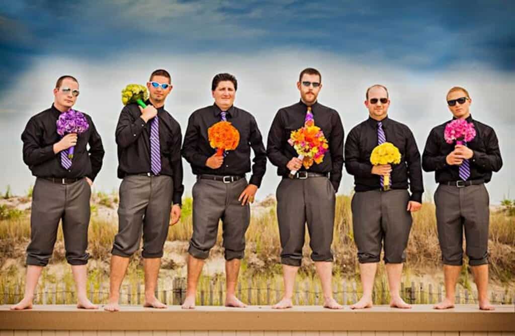 Six barefoot men in matching black shirts, gray pants, and striped purple ties stand near a sand dune, holding colorful bouquets.