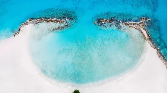 An aerial view shows a circular sandy beach with clear blue water, bordered by two curved rocky formations.